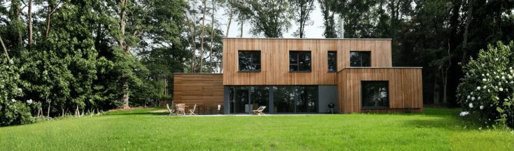 Maison moderne à deux étages avec un extérieur en bois, de grandes fenêtres et une pelouse spacieuse, entourée de grands arbres et de verdure. Des chaises d'extérieur et une table sont disposées sur le patio.