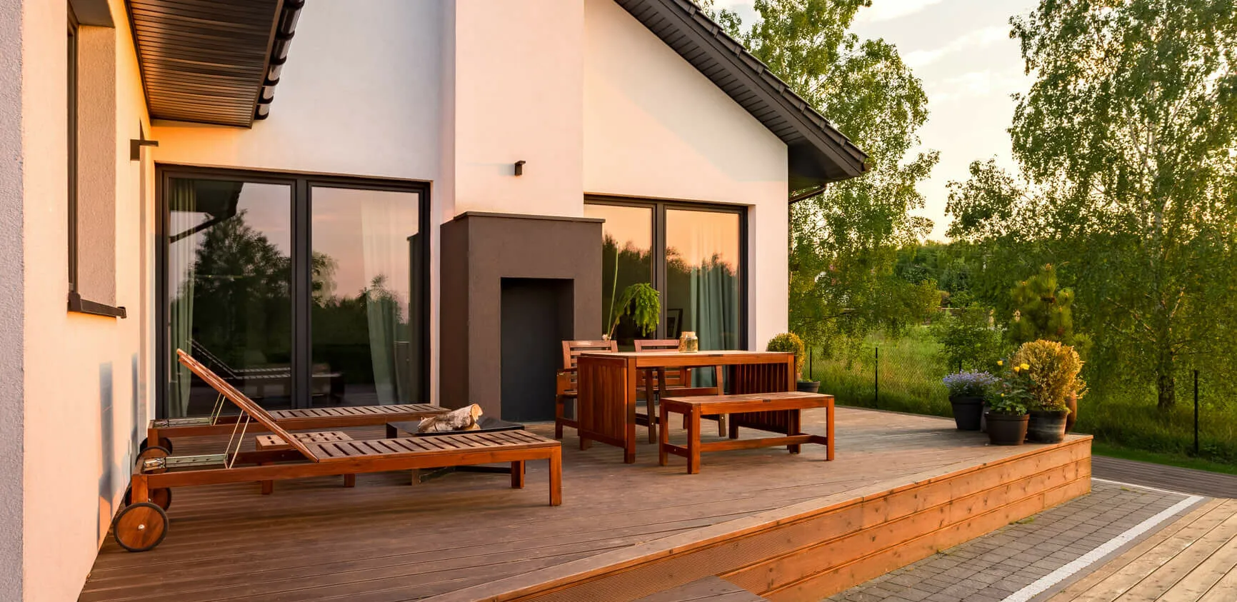 Une maison moderne aux murs blancs et au toit sombre dispose dune spacieuse terrasse en bois, encadrée par des fenêtres à châssis épurées. La terrasse est équipée de chaises longues, dune table à manger et de plantes en pot. Le soleil couchant projette une lueur chaleureuse et des arbres sont visibles en arrière-plan.