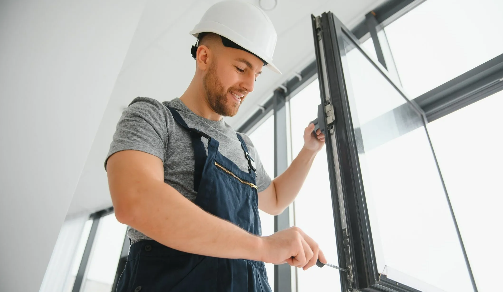 Un homme portant un casque de chantier blanc et une salopette travaille sur une fenêtre. Il utilise un tournevis pour faire des ajustements. La pièce est lumineuse avec de grandes fenêtres en arrière-plan.