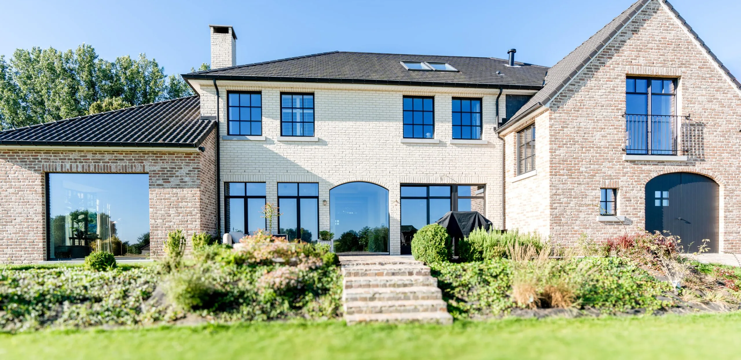 Une grande maison en brique à deux étages avec un toit noir, de multiples fenêtres et une cheminée. La cour avant comprend un jardin soigneusement aménagé avec des marches menant à l’entrée. Le ciel est clair et bleu.