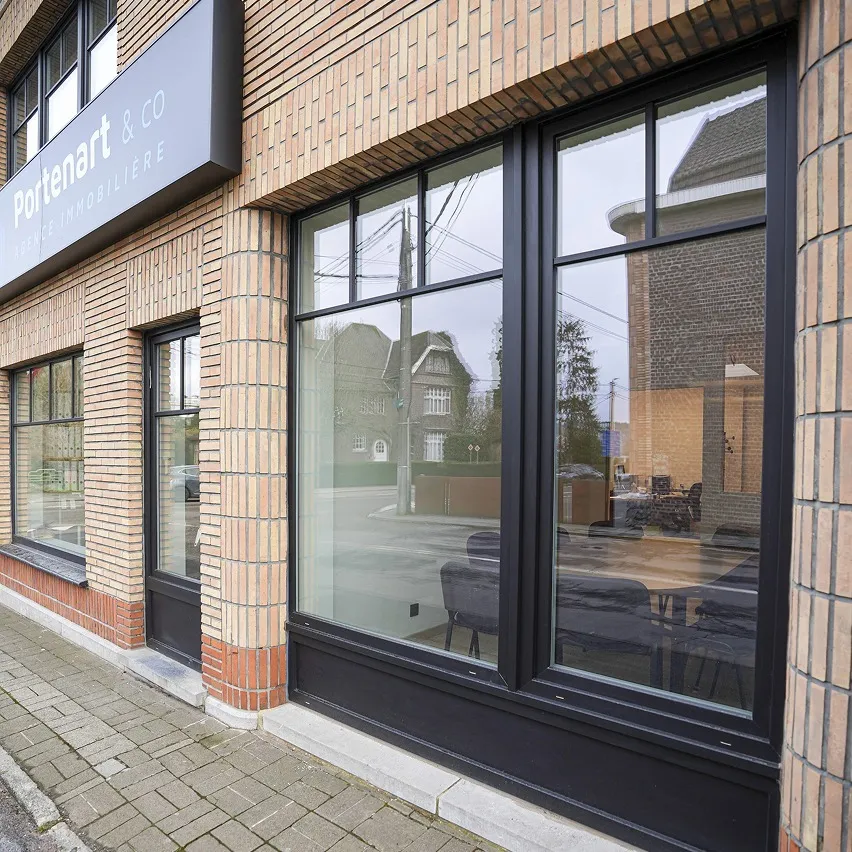 Extérieur d'un immeuble en brique avec une grande fenêtre laissant entrevoir le reflet de la rue et des maisons voisines. Au-dessus, un panneau indique « Portenart & Co Vente immobilière ». Un trottoir pavé longe le bâtiment.
