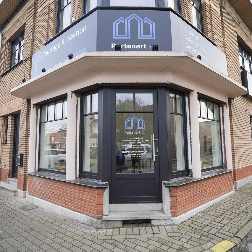 Vue de la rue d'un immeuble de bureaux d'angle avec de grandes fenêtres et une porte. Le panneau au-dessus indique « Portenart & Cie » avec un texte supplémentaire en dessous. L'entrée comporte un logo et un texte sur la porte vitrée. L'extérieur est fait de briques et de ciment.