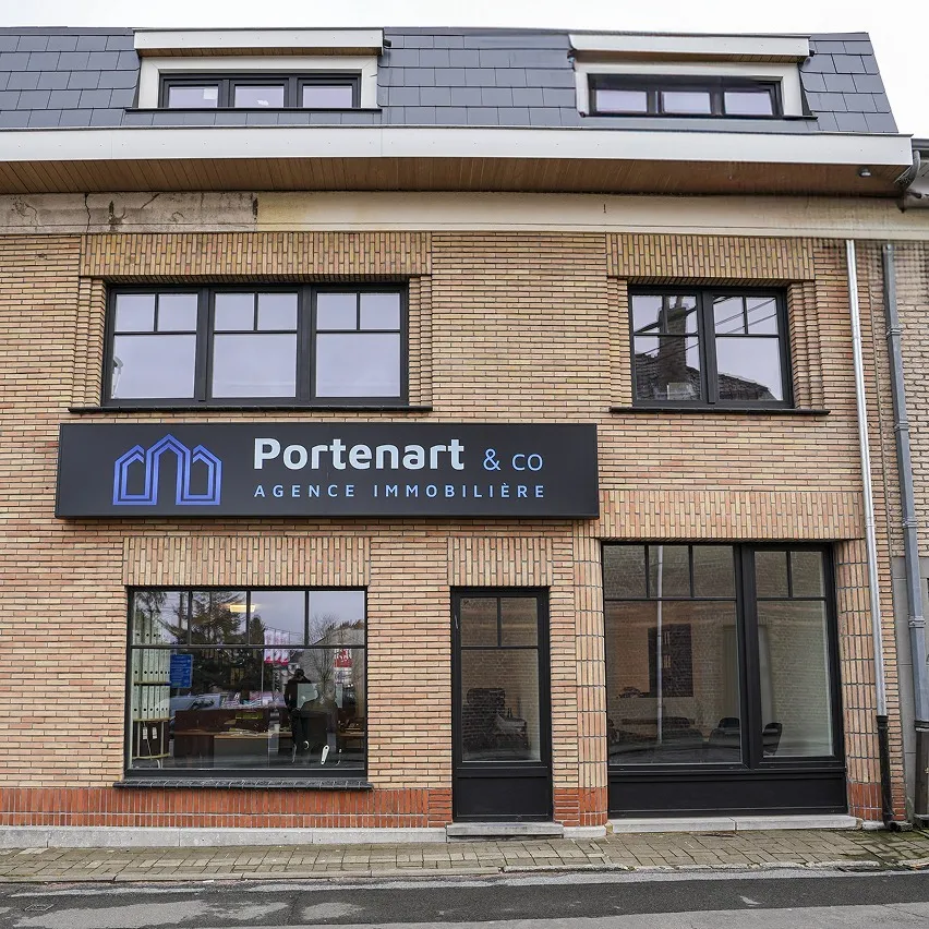 Un bâtiment de deux étages avec une façade en briques abrite l'agence immobilière « Portenart & Co Agence Immobilière ». Une grande enseigne noire avec des graphiques et du texte bleus est affichée au-dessus des portes et fenêtres en verre du rez-de-chaussée. Les étages supérieurs disposent de fenêtres supplémentaires.