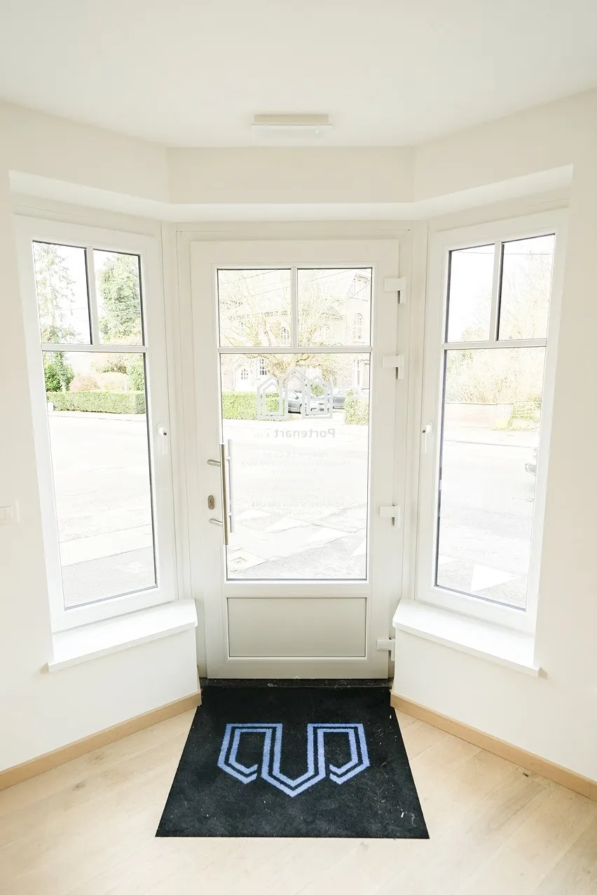 Une porte d'entrée avec une porte vitrée et deux hautes fenêtres latérales. À l'extérieur, on peut voir des arbres et un bâtiment à travers la vitre. À l'intérieur, un tapis noir avec un logo bleu et blanc est posé sur le sol en bois.