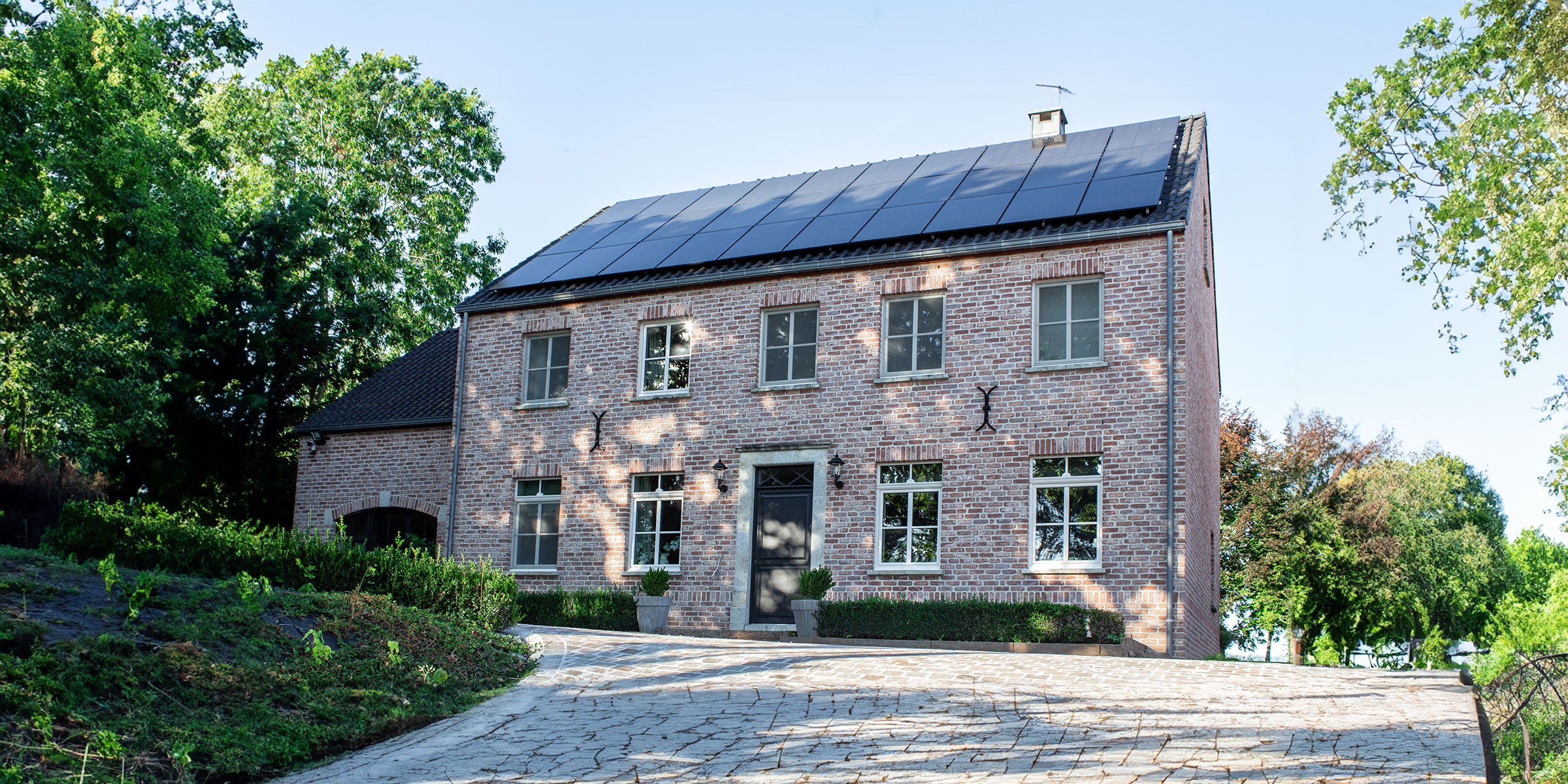 Une maison en brique avec plusieurs fenêtres et une porte noire, située sur une allée pavée. Des panneaux solaires sont installés sur le toit. La maison est entourée darbres et de verdure sous un ciel bleu clair.