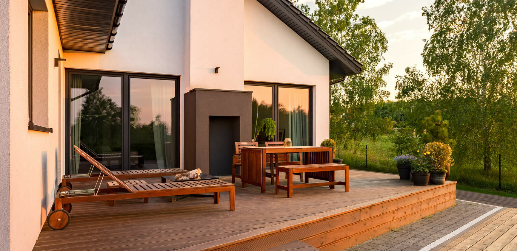 Une maison moderne aux murs blancs et au toit sombre dispose dune spacieuse terrasse en bois, encadrée par des fenêtres à châssis épurées. La terrasse est équipée de chaises longues, dune table à manger et de plantes en pot. Le soleil couchant projette une lueur chaleureuse et des arbres sont visibles en arrière-plan.