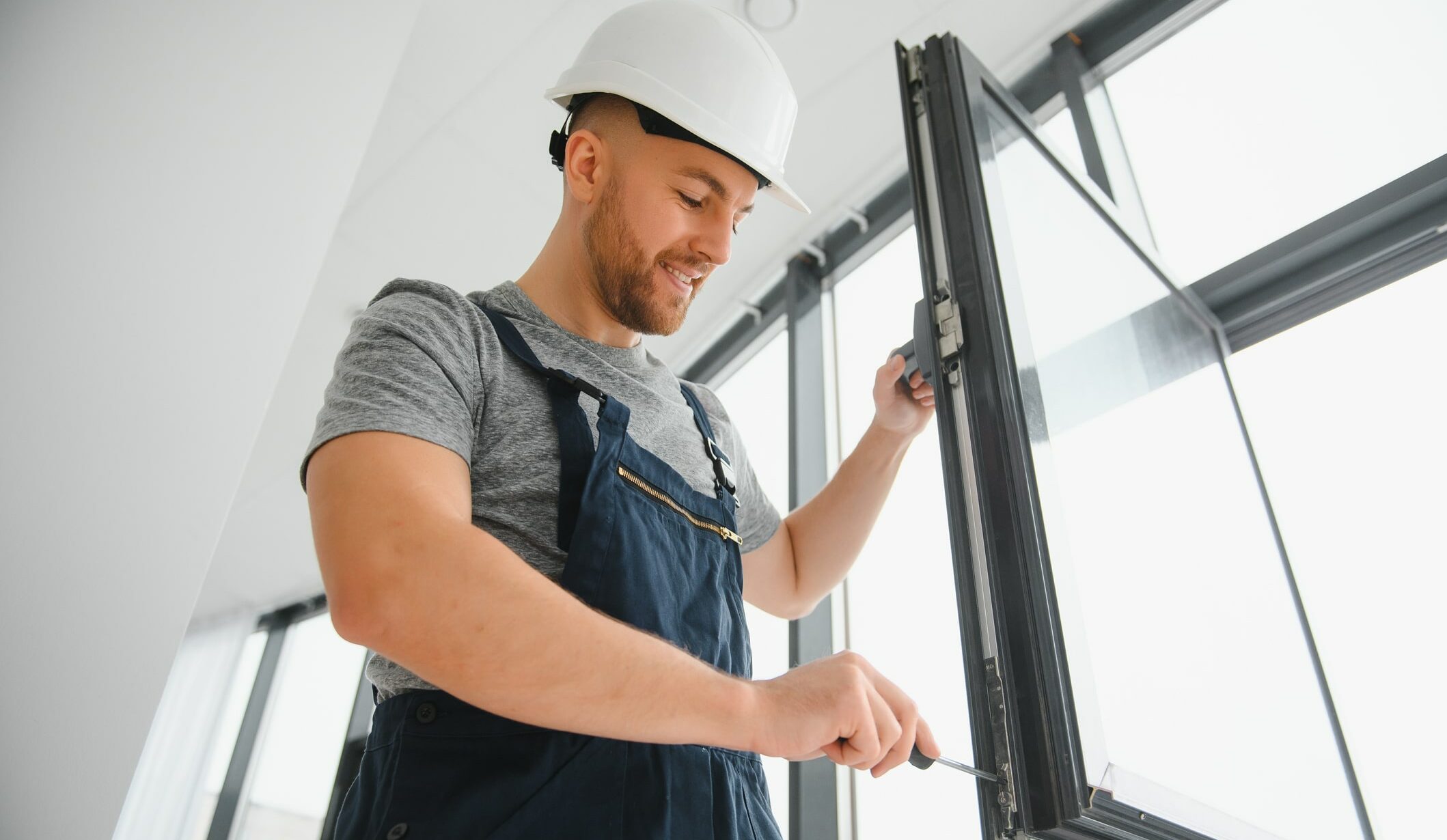 Un homme portant un casque de chantier blanc et une salopette travaille sur une fenêtre. Il utilise un tournevis pour faire des ajustements. La pièce est lumineuse avec de grandes fenêtres en arrière-plan.