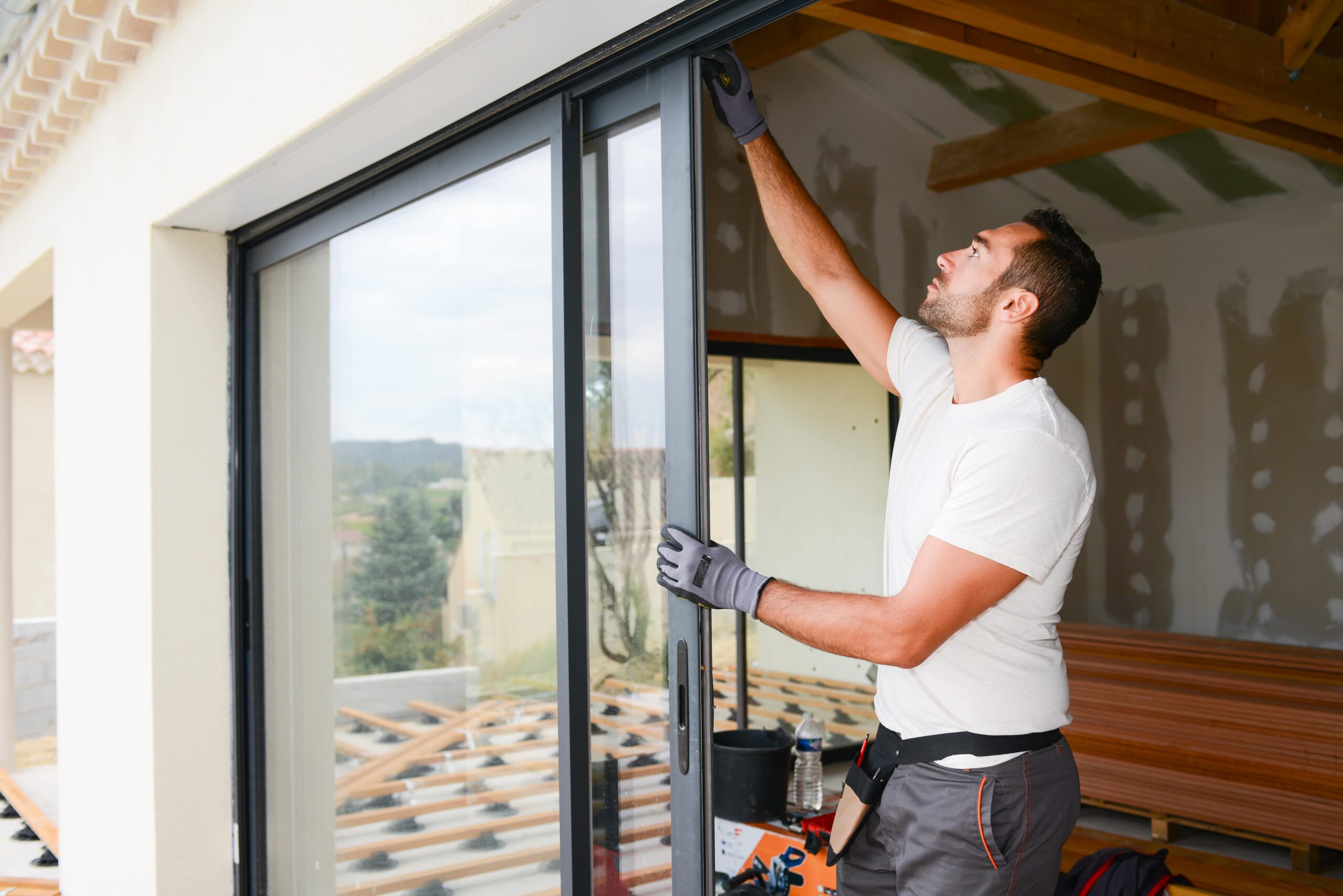 Un homme portant des gants installe ou ajuste une grande porte coulissante en verre dans une maison en construction, avec des murs non finis et des poutres de plafond apparentes visibles à l'arrière-plan.